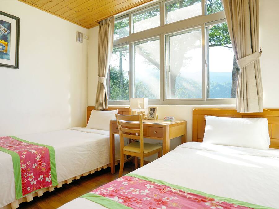 A room with two beds, a desk, and a chair. There is a window with curtains, and a painting on the wall.