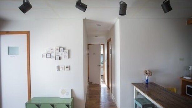 A hallway with a wooden floor, a green bench, and a table with a vase. There are multiple light fixtures on the ceiling. - Lucidbonbon - Hostel (Seogwipo)