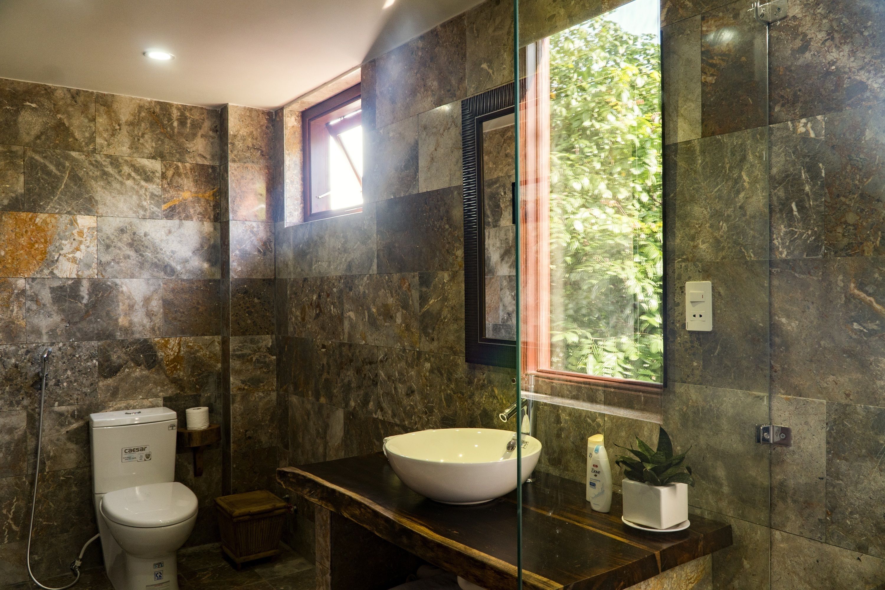 A bathroom with a marble wall, a white sink, a toilet, and a window with a view of greenery.