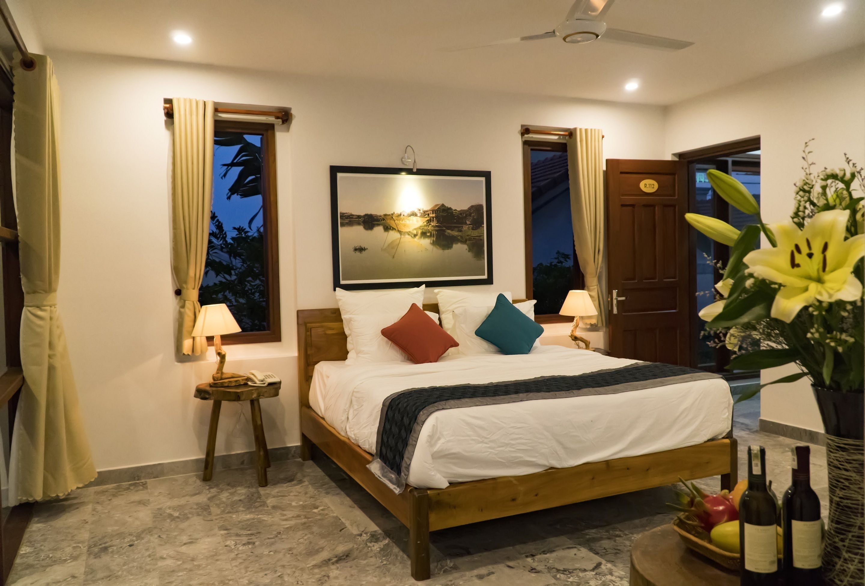 A bedroom with a bed, bedside lamps, a nightstand, a framed picture on the wall, a door, and a potted plant.