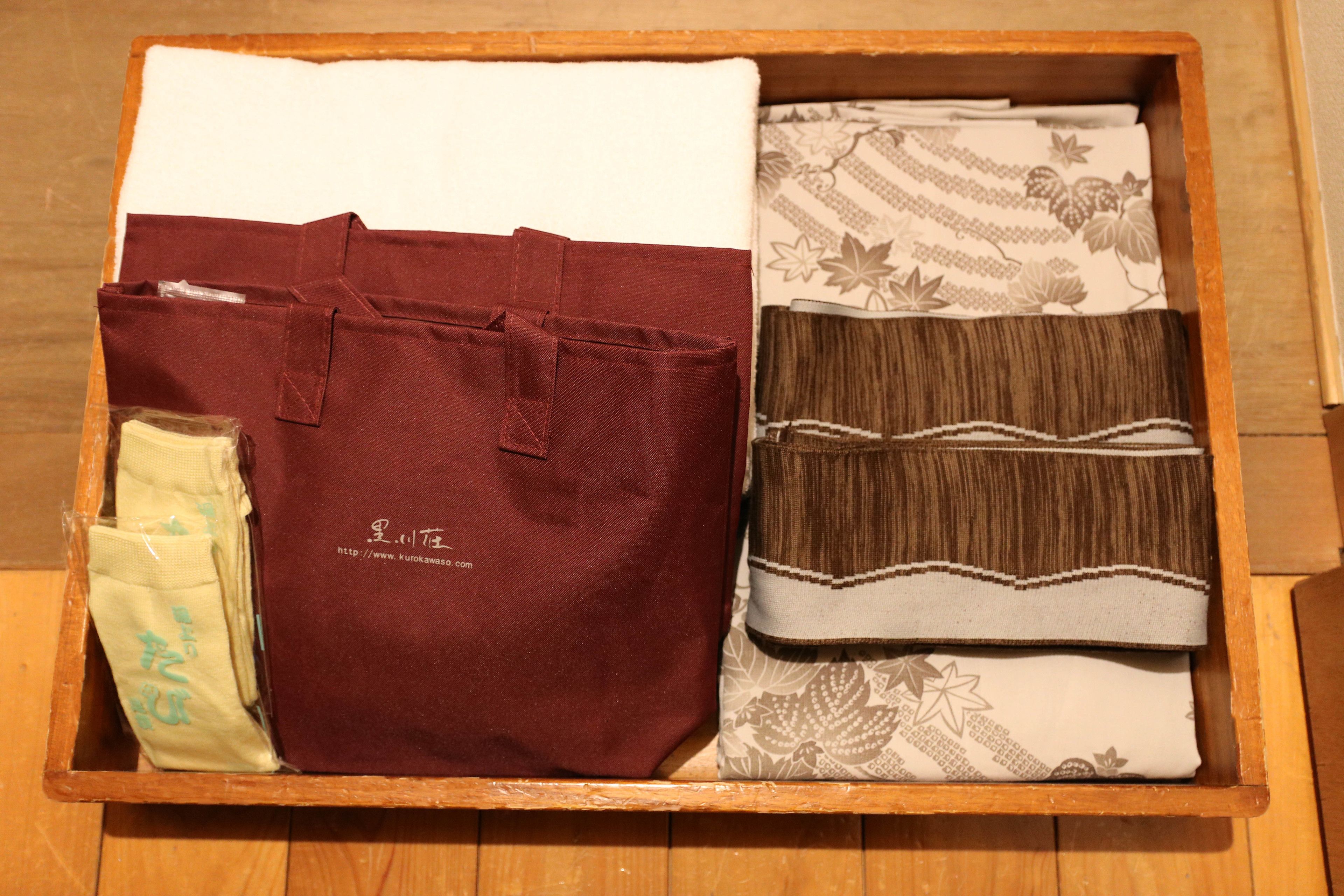 Una caja de madera con un bolso color burdeos, un paño estampado y un trozo de tela marrón doblado.