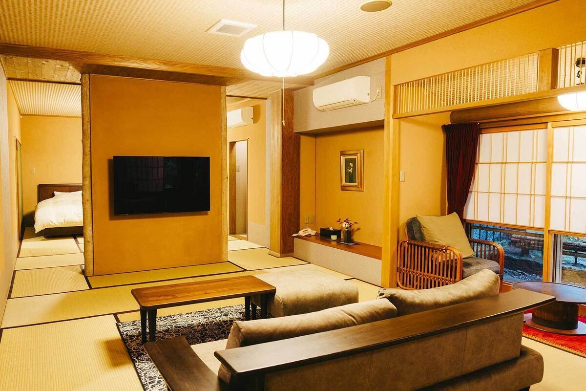 A traditional Japanese-style room with tatami flooring, a sofa, a coffee table, a television, and a view of a bridge through a shoji screen.