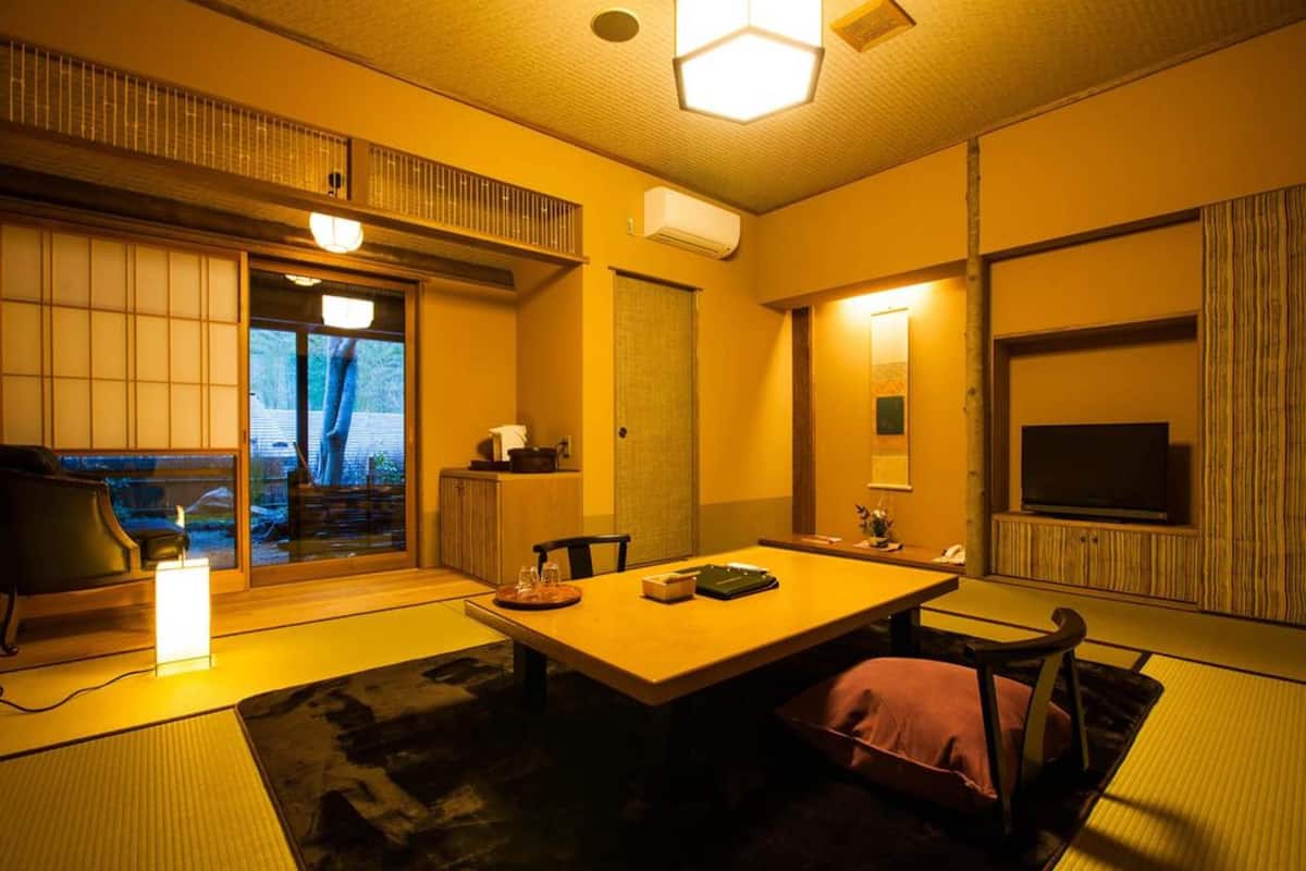 A traditional Japanese-style room with tatami flooring, a low table with cushions, a sliding door leading to a balcony, and a television on a wooden shelf.
