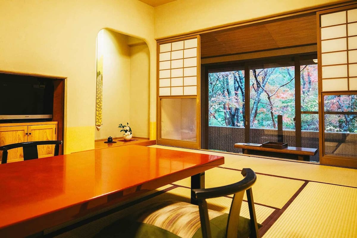 A traditional Japanese room with tatami flooring, a wooden table, a television, and a view of trees through a shoji screen.
