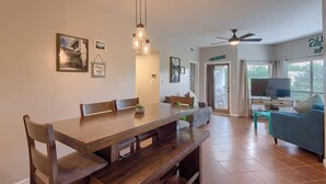 Un comedor con mesa y sillas de madera, un banco, ventilador de techo y televisor en una sala de estar con sofá y cortinas.