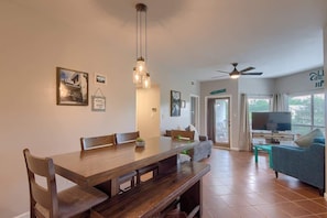A dining area with a wooden table and chairs, a bench, a ceiling fan, and a television in a living room with a sofa and curtains. - IC 302 Ripley River Haus (New Braunfels)