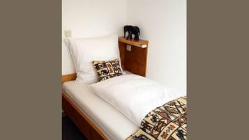 A single bed with white bedding, a patterned pillow, and a small shelf with a figurine.