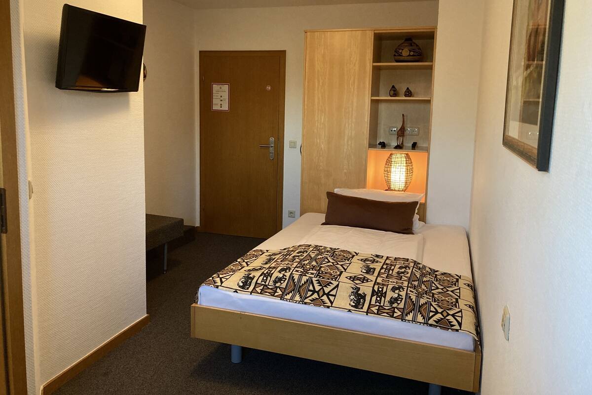 Une petite chambre à coucher, bien rangée, avec un lit, une table de chevet, une lampe et un téléviseur fixé au mur.