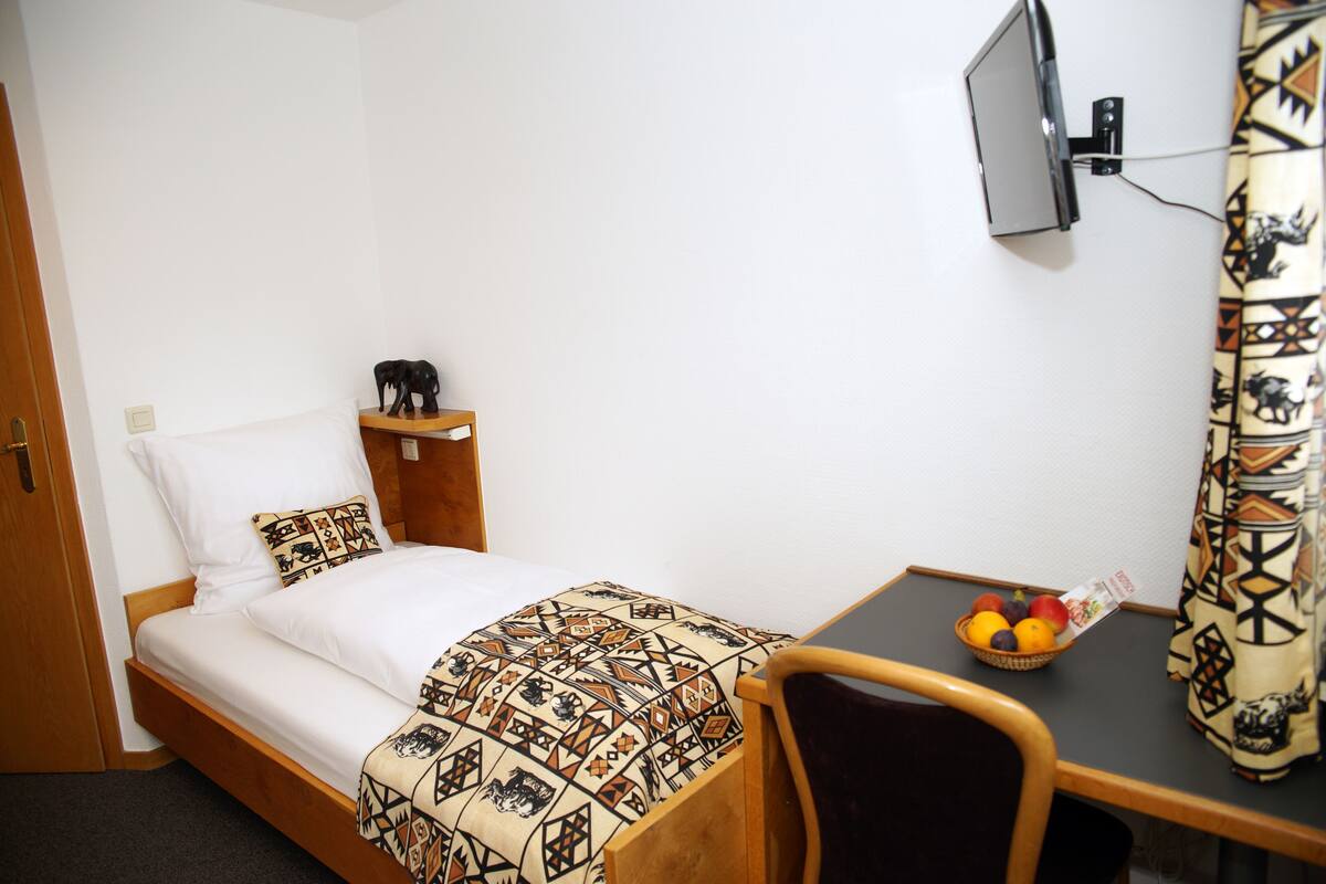 Une chambre à coucher avec un lit, une table de chevet sur laquelle se trouve un panier de fruits, un téléviseur fixé au mur et un rideau à motifs.