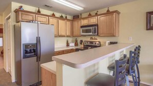 Une cuisine avec des meubles en bois, un réfrigérateur, un four à micro-ondes, un îlot central avec des chaises et un panneau indiquant le mot « Mecha ».