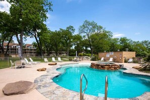 A swimming pool with lounge chairs, a stone deck, and a building in the background. - WW D301 On River Time (New Braunfels)