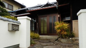 Un edificio japonés tradicional con tejado de tejas, puerta de madera con paneles de vidrio y un pequeño jardín con plantas y un sendero de piedra que lleva a la entrada.
