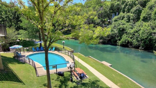A swimming pool with a wooden deck, surrounded by grass and trees, with a view of a river.
