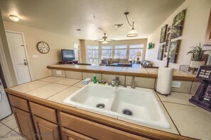 A kitchen with a double sink, wooden cabinets, a ceiling fan, and a window overlooking a deck.