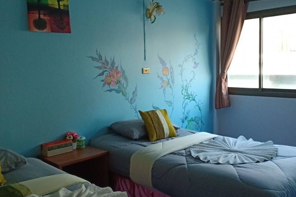 A bedroom with two beds, a nightstand, a wall with floral and tree paintings, and a window with curtains.