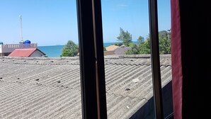Une vue à travers une fenêtre donnant sur l’extérieur d’un bâtiment, avec un toit en tôles ondulées, et une vue lointaine de la mer et d’une grue.
