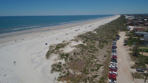 An aerial view of a beach with a row of parked cars along the shore, a sandy beach, and a clear sky.