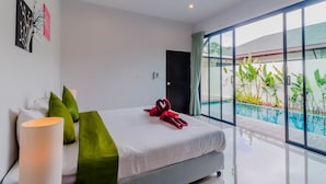 Quarto de hotel moderno com cama grande, almofadas verdes, cabeceira branca e vista para uma piscina através de janelas do chão ao teto.