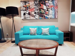 A teal sofa with two decorative pillows, a glass coffee table, and a colorful abstract painting on the wall. - Greystone Haus at D' Majestic Place (Kuala Lumpur)