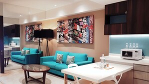 A modern living room with a teal sofa, a glass coffee table, and a white dining table. There is a large abstract painting on the wall and a microwave on the counter. - Greystone Haus at D' Majestic Place (Kuala Lumpur)
