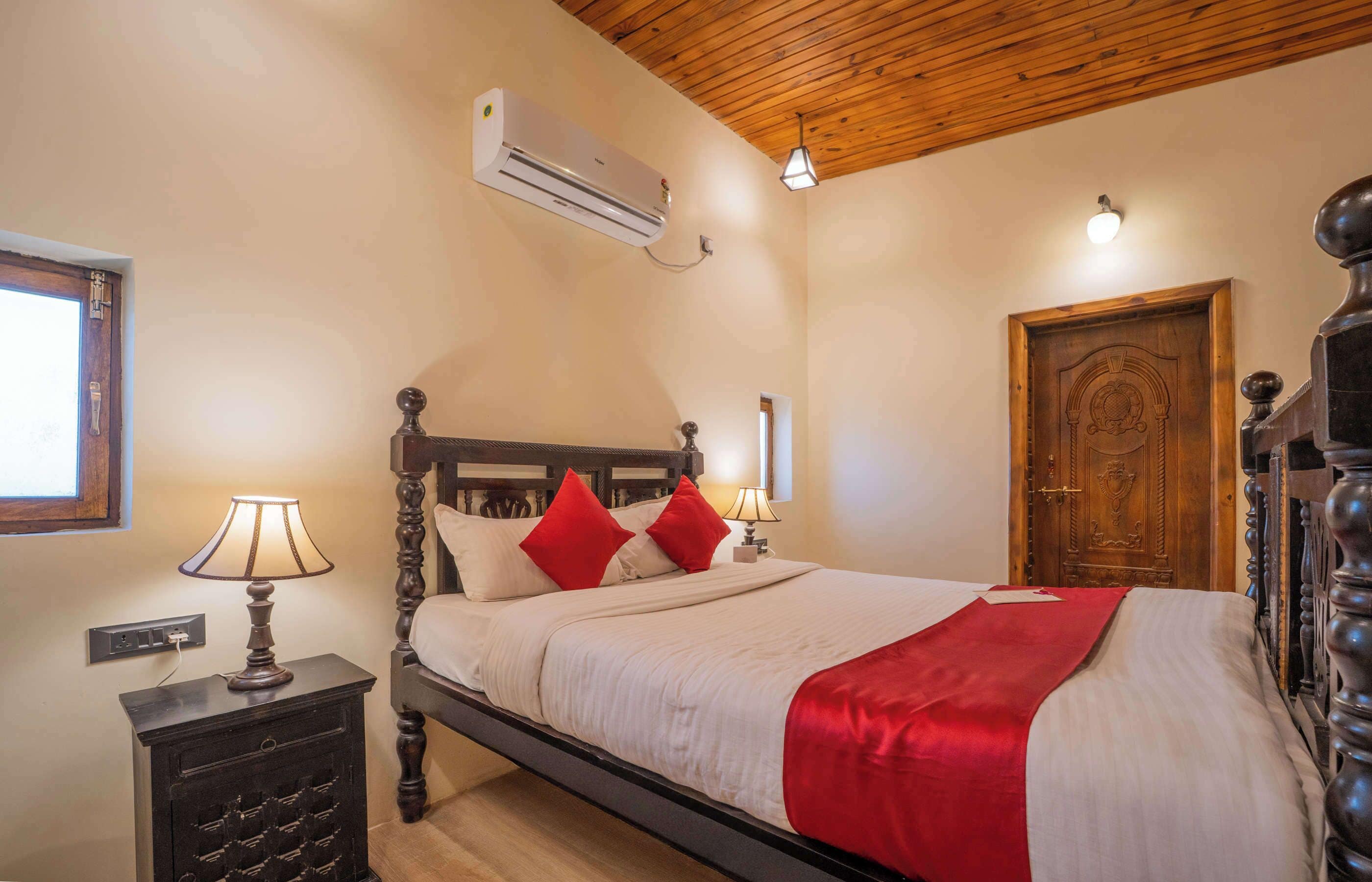 A bedroom with a wooden ceiling, a bed with a red throw, a nightstand with a lamp, and a wooden door.