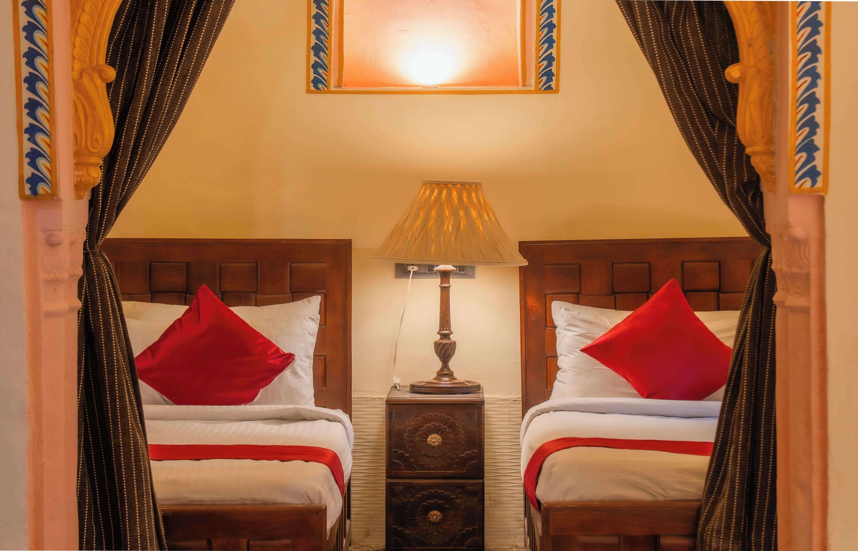 A room with two beds, each with a red pillow, a wooden nightstand with a lamp, and a mirror above the beds.