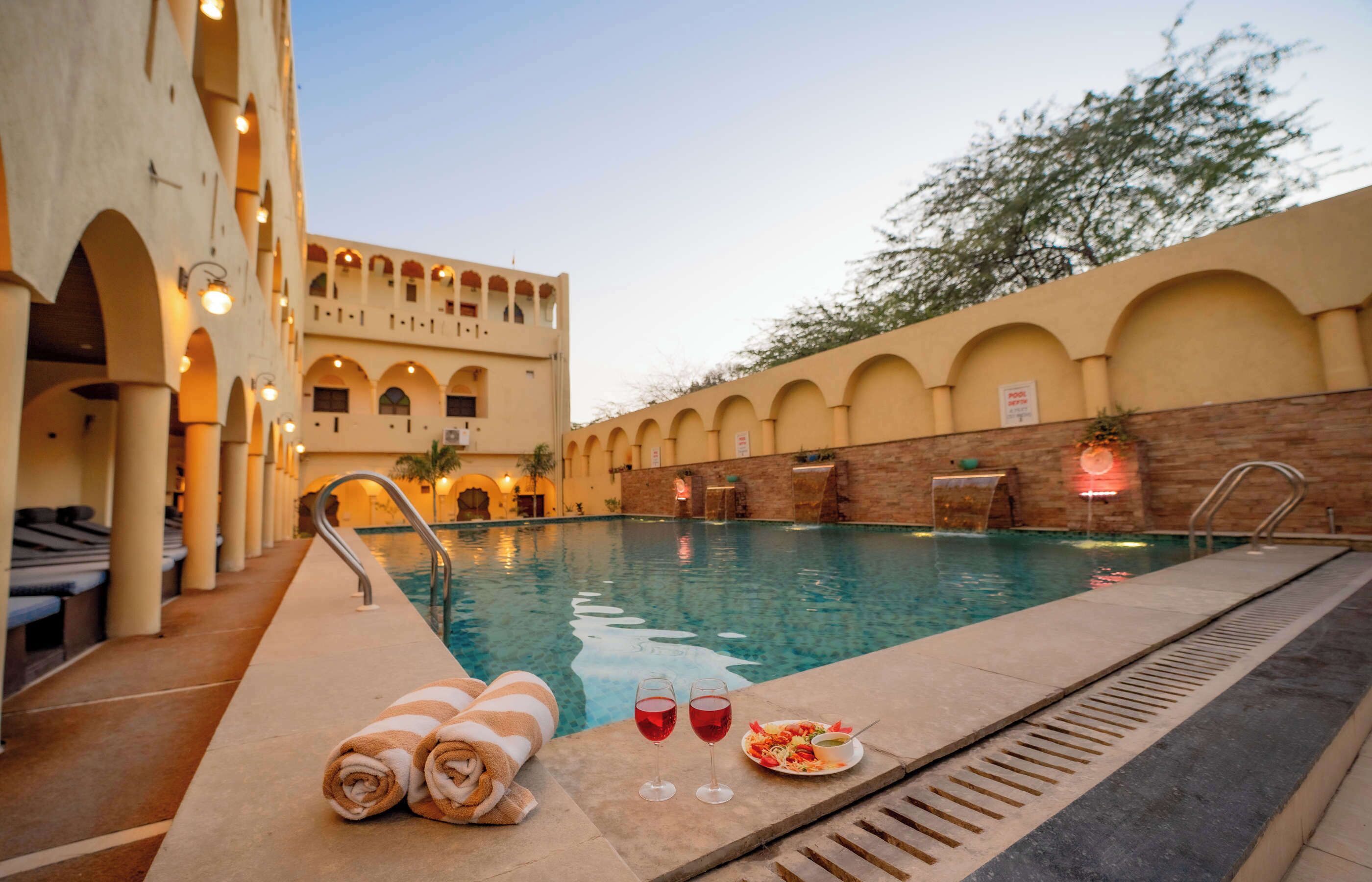 A swimming pool with a sunken seating area, a poolside dining setup, and a building with arched windows and columns.