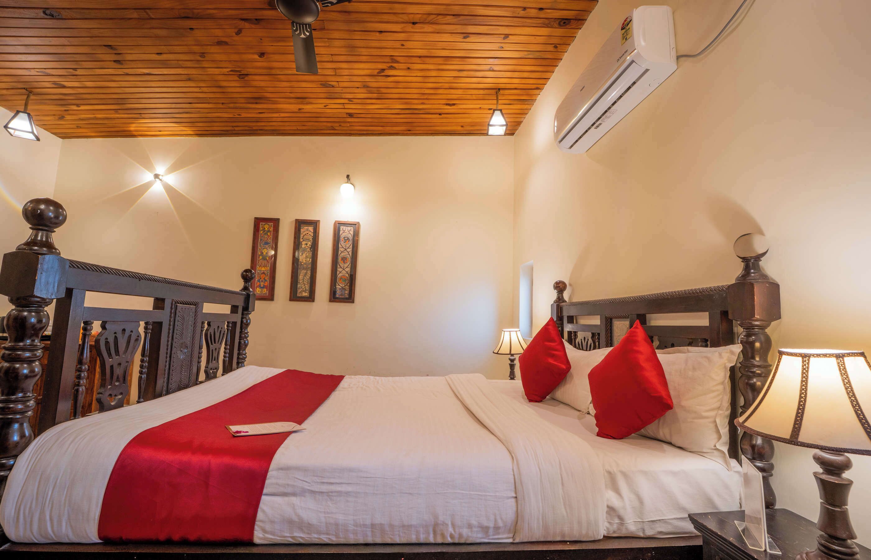 A hotel room with a wooden ceiling, a bed with white and red bedding, a nightstand with a lamp, and wall-mounted air conditioning.