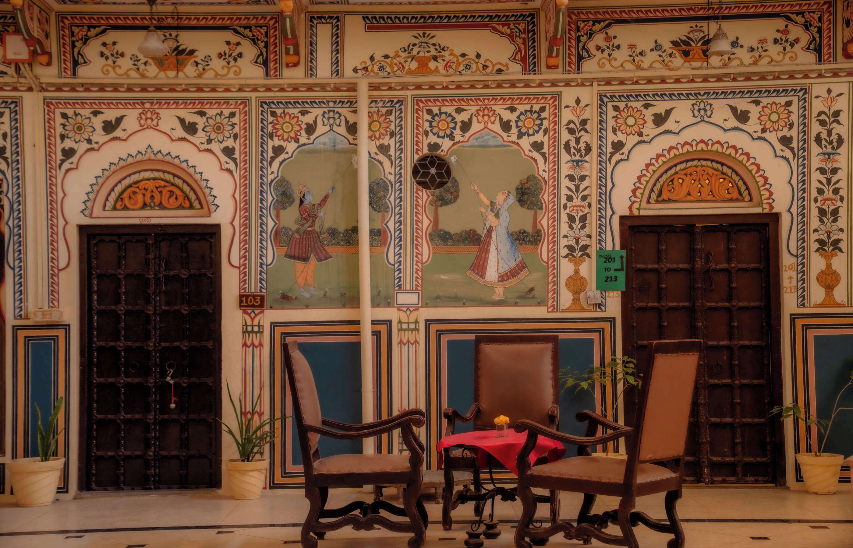 Une pièce dont les murs sont ornés de décorations raffinées, avec deux chaises en bois, une table recouverte d’une nappe rouge et une porte portant un numéro.