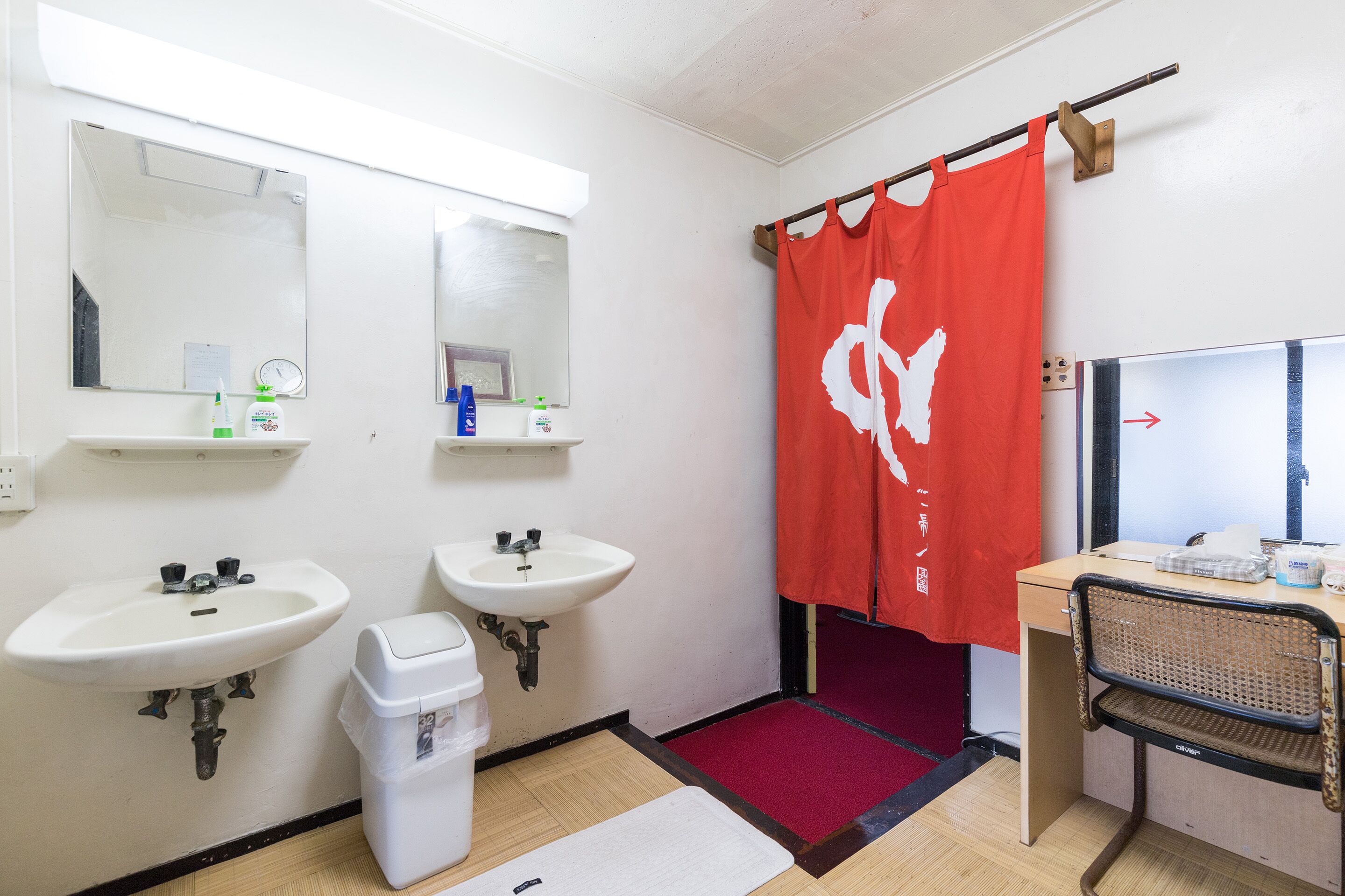 A bathroom with two sinks, a mirror, a shower curtain, a trash bin, and a desk with a chair.