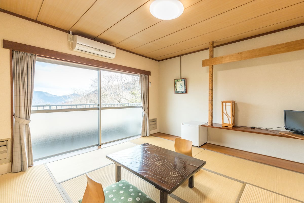 Una habitación con suelo de tatami, una mesa de madera, un televisor y vistas a montañas a través de un ventanal amplio.