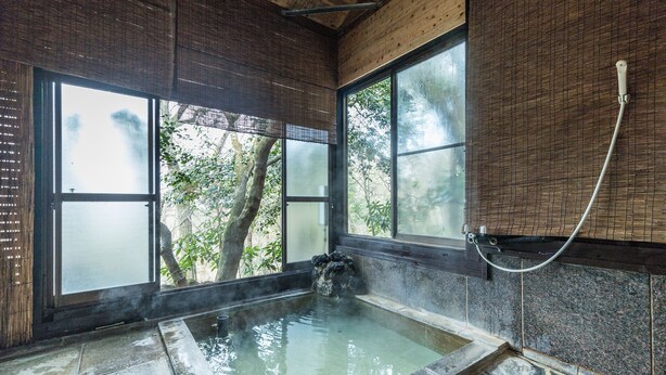 Un onsen japonés tradicional con un baño de piedra, persianas de bambú y vistas a árboles.