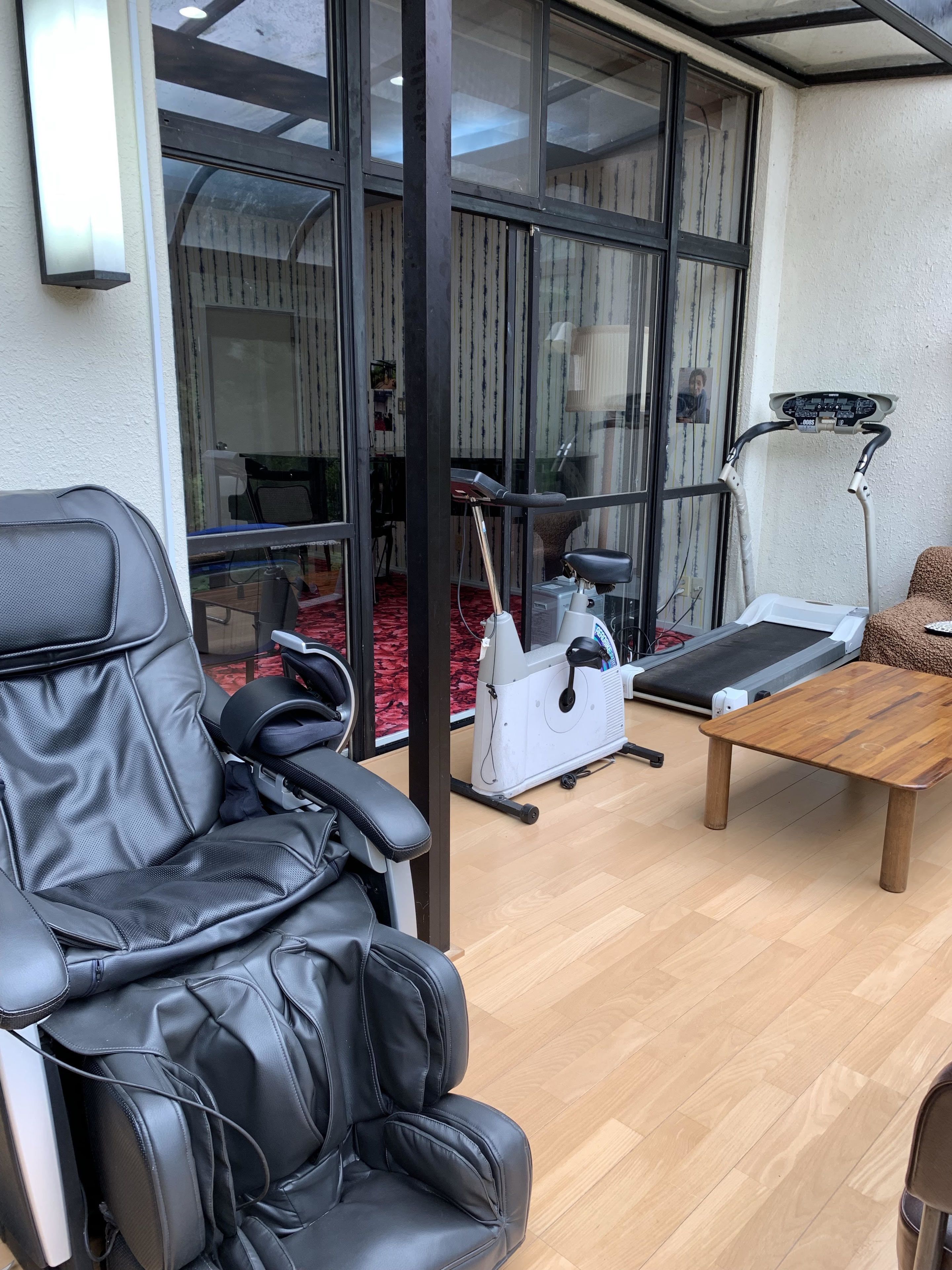 A massage chair, a treadmill, and a wooden table in a room with large windows and a red carpet.
