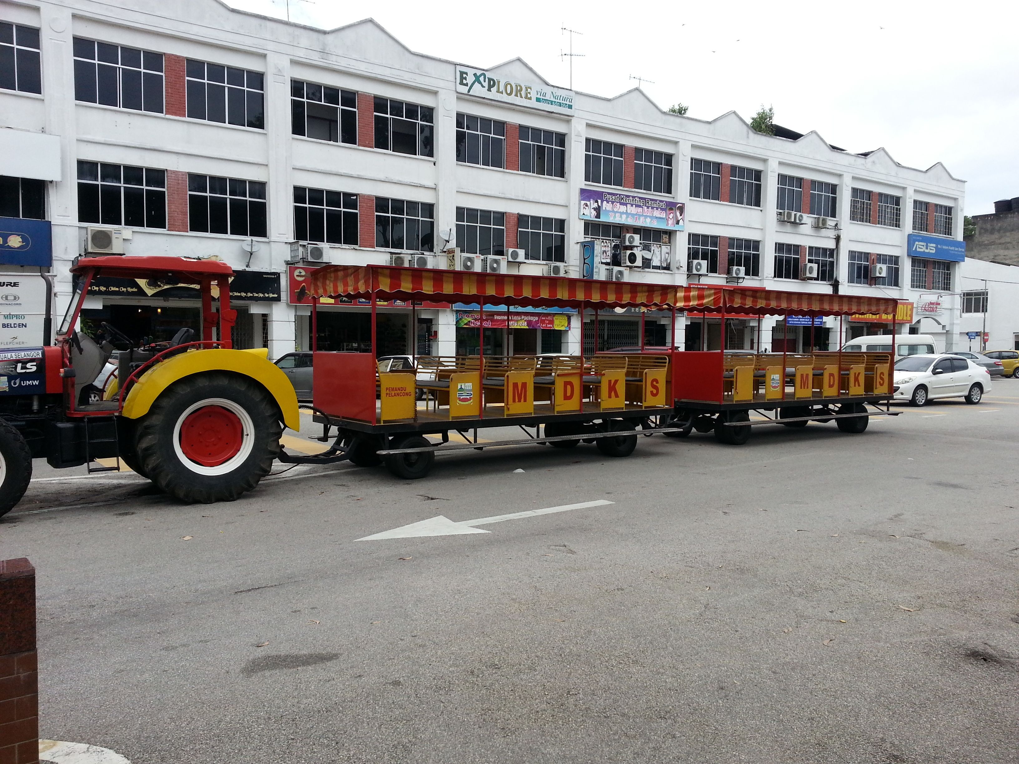 A stationary train with open carriages and a tractor pulling it, parked in front of a building with multiple signs and windows.