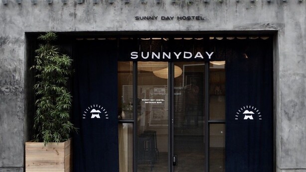 A hostel with a dark blue entrance, a sign reading "SUNNYDAY HOSTEL," and a small plant to the left. - Sunny Day Hostel (Takamatsu)