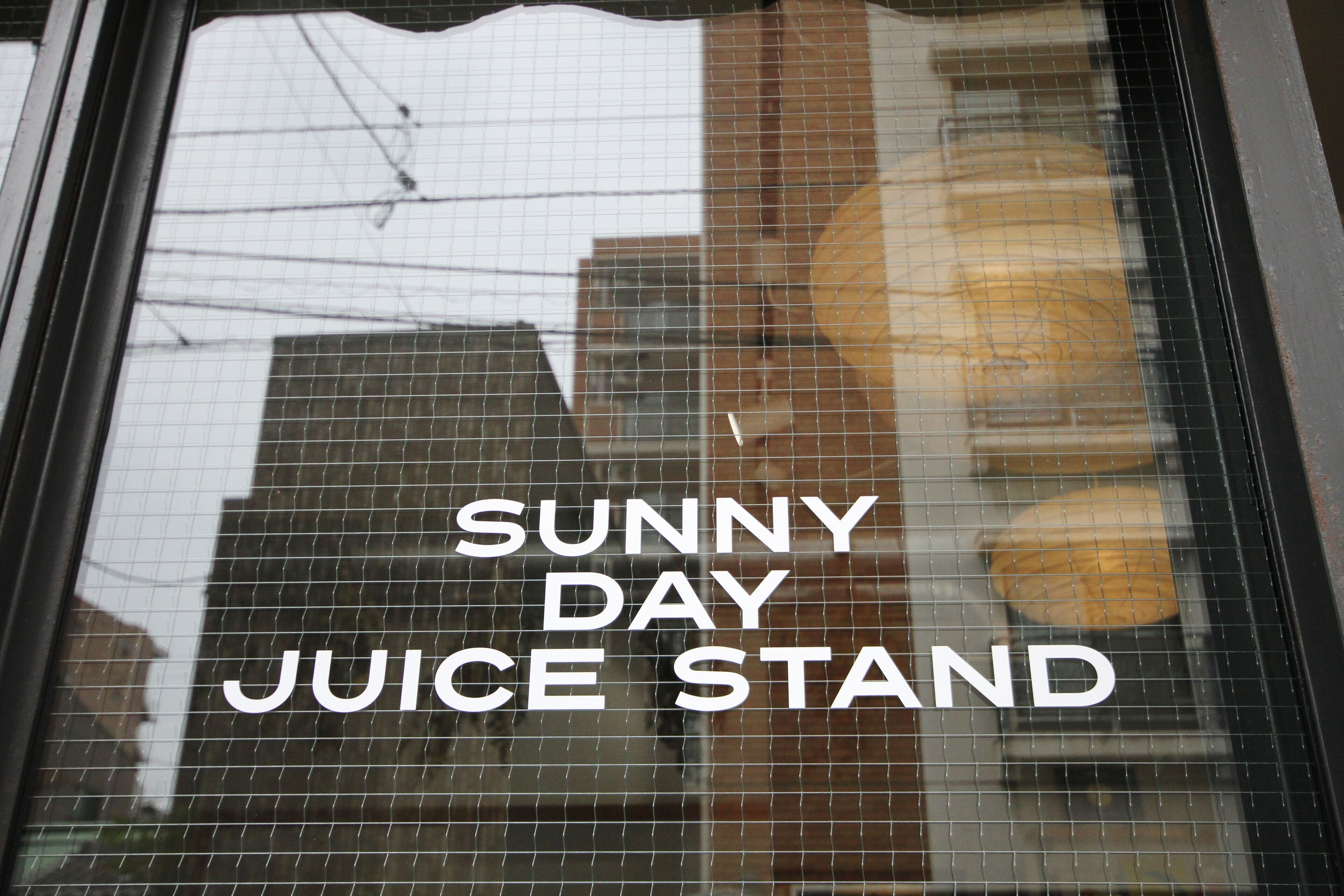 Janela exibindo o texto "SUNNY DAY JUICE STAND" e reflexos de edifícios e fios de energia.