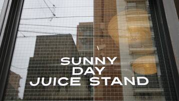 Janela exibindo o texto "SUNNY DAY JUICE STAND" e reflexos de edifícios e fios de energia.