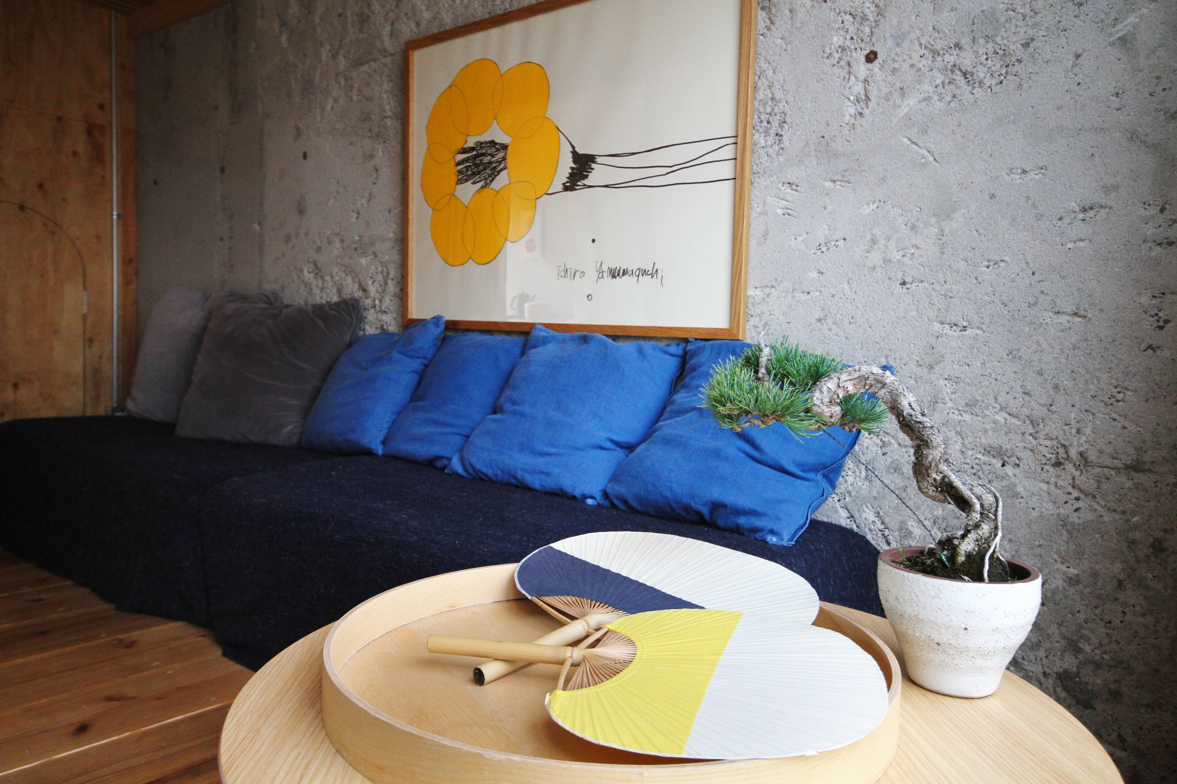 Ein Zimmer mit einem Bett, blauen Kissen, einem gerahmten Kunstwerk, einem Bonsai-Baum und einem runden Tisch mit Ventilatoren und einer Schüssel.