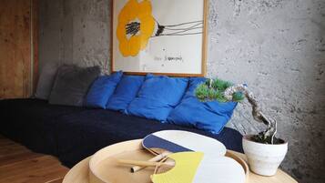Ein Zimmer mit einem Bett, blauen Kissen, einem gerahmten Kunstwerk, einem Bonsai-Baum und einem runden Tisch mit Ventilatoren und einer Schüssel.