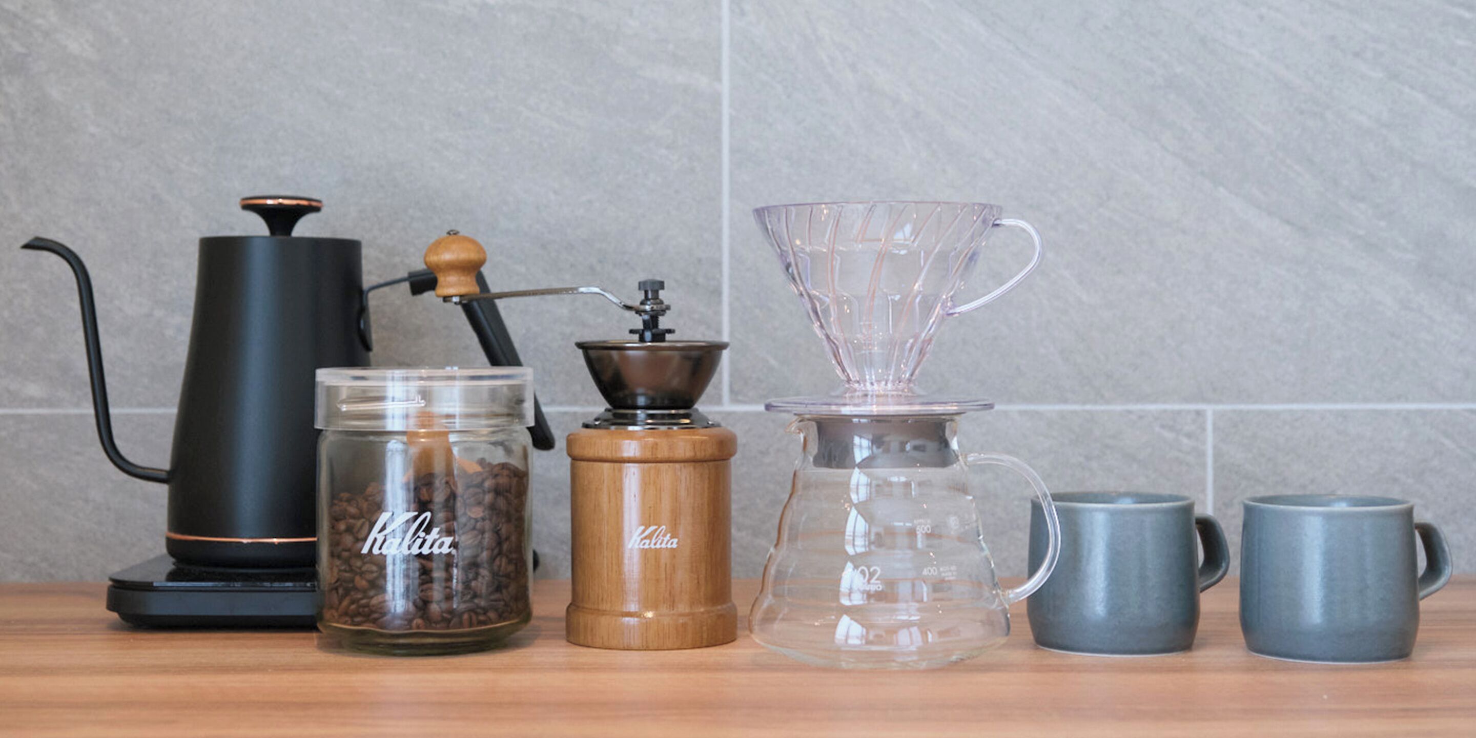 A coffee maker, a grinder, a glass carafe, and two mugs on a wooden surface.