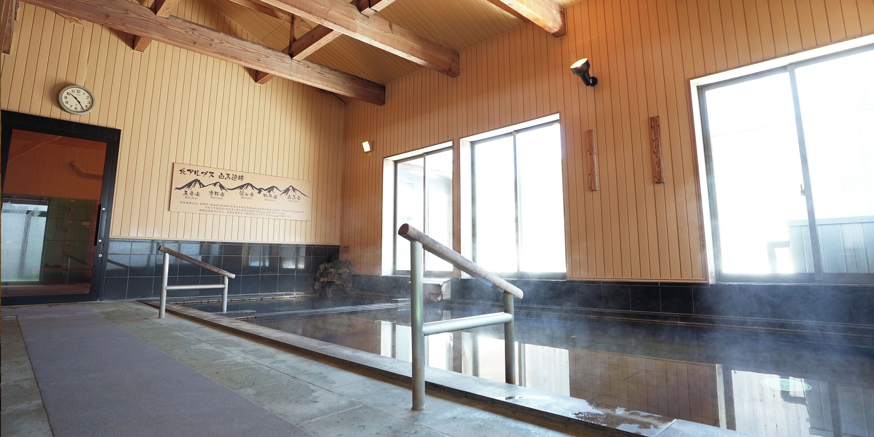 A traditional Japanese onsen with wooden paneling, a stone hot spring, and a wooden staircase leading to the entrance.