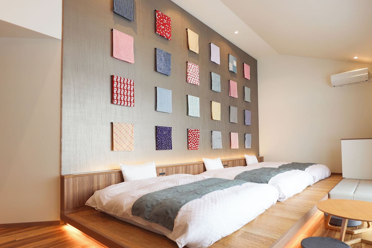 A modern hotel room with two beds, a sofa, a small table, and a wall decorated with various patterned fabrics.