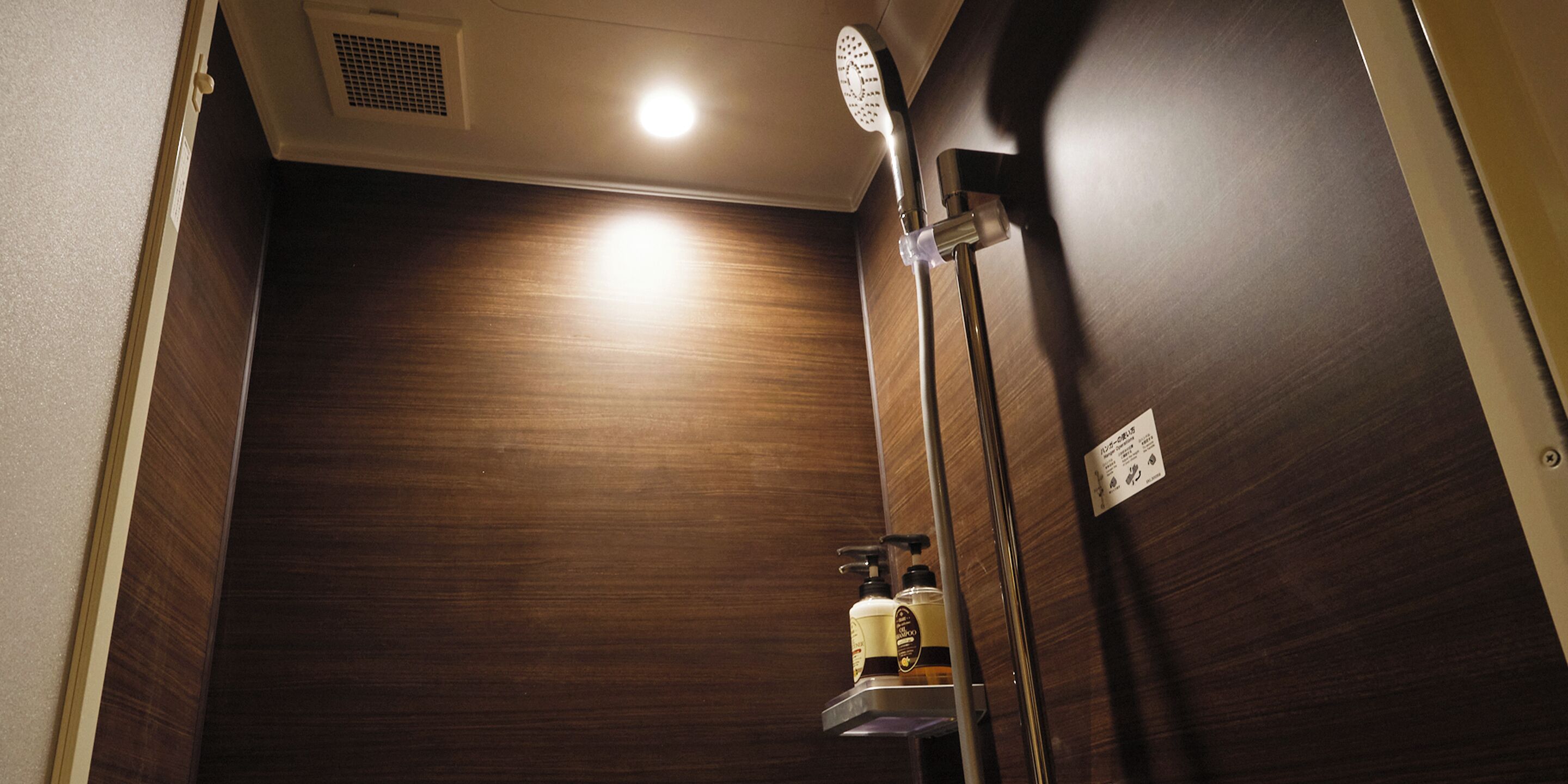 A wooden shower stall with a handheld showerhead, a soap dispenser, and a light fixture on the ceiling.
