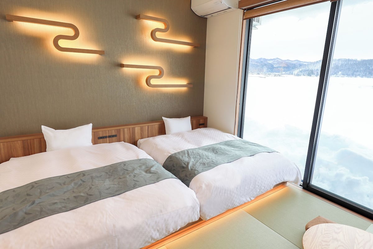 A hotel room with two beds, a large window with a view of mountains, and a wall-mounted light fixture.