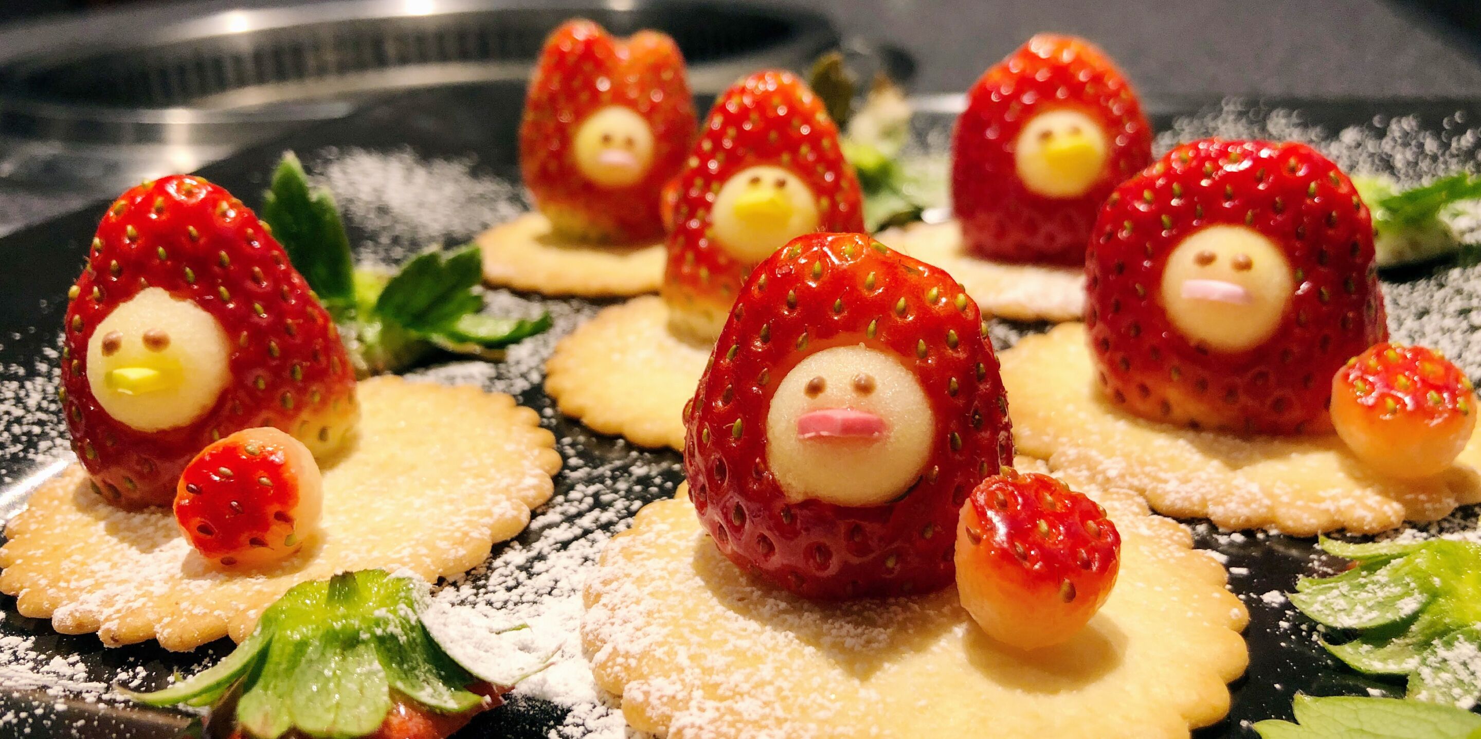 Fresas con caras en un plato, acompañadas de galletas pequeñas y hojas de menta.
