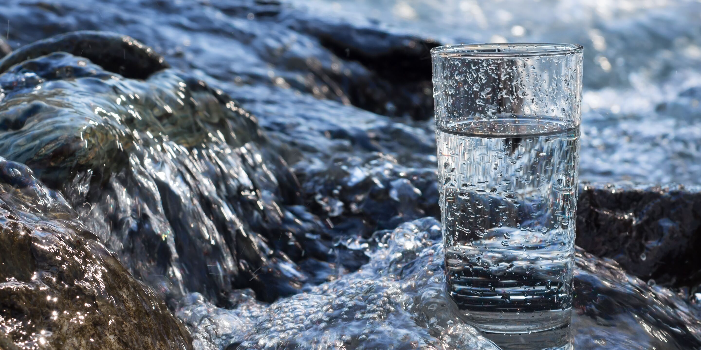 A glass of water placed on a rocky surface with flowing water in the background.