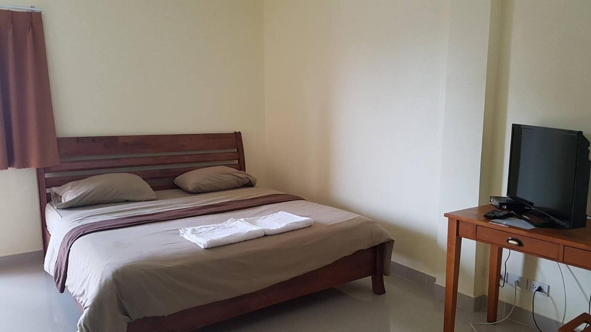Una cama individual con cabecera de madera, una mesita de noche con televisor y control remoto, y un enchufe en la pared.
