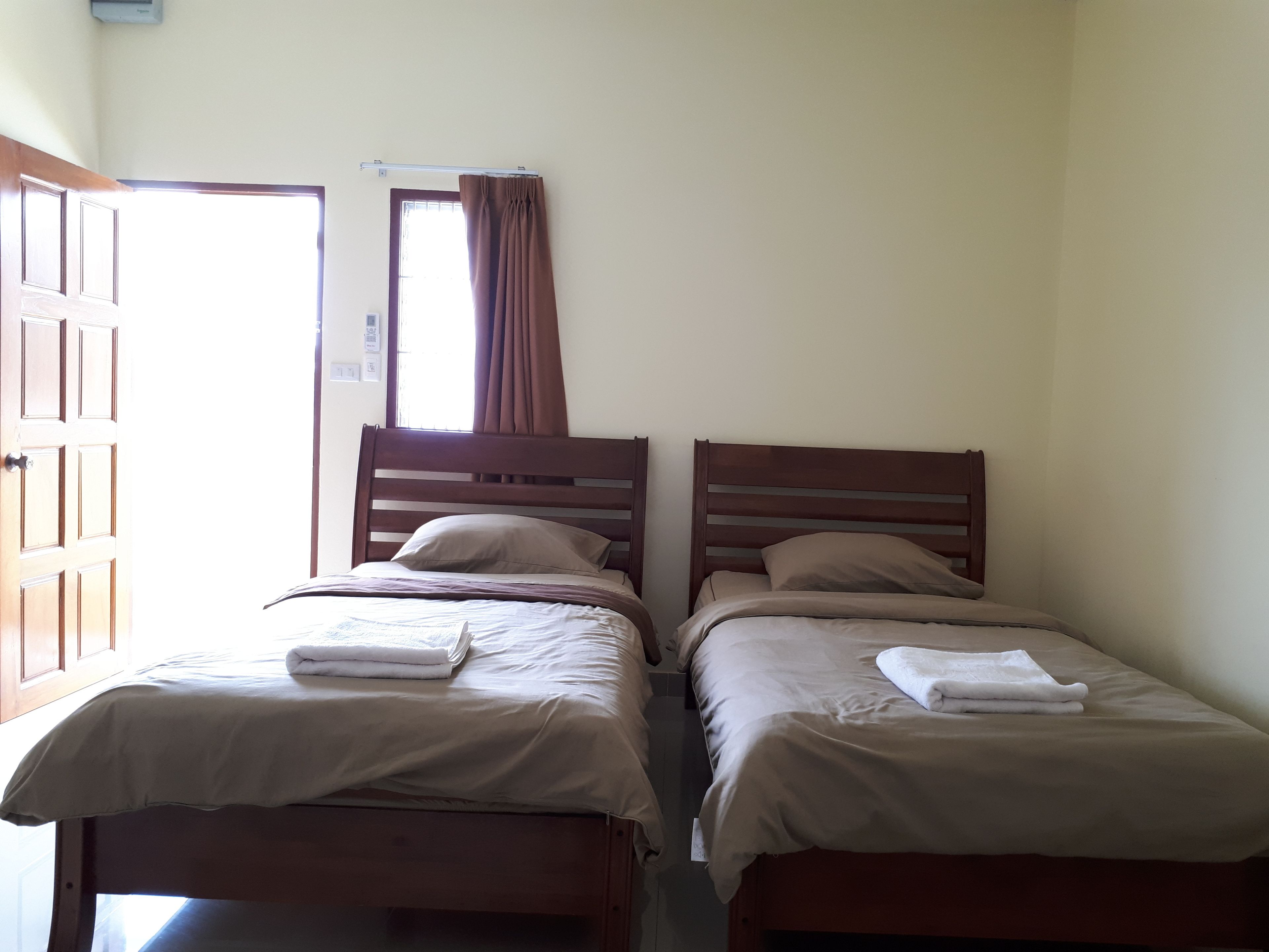 A room with two wooden beds, each with a pillow and a folded towel. There is a window with curtains, and a door is visible on the left side.
