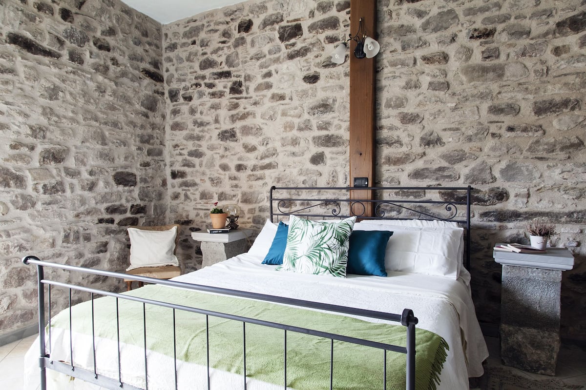 A bedroom with a stone wall, a metal bed frame, and a nightstand with a lamp.