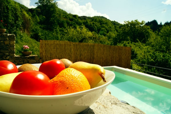 A bowl of fruit on a balcony with a pool and a view of a lush green hillside.